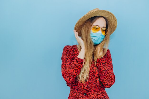 Una ragazza emotiva in un abito rosso, cappello e una maschera medica su sfondo blu. Viaggio annullato a causa dell'epidemia di coronavirus.