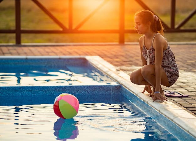 Una ragazza felice abbronzata grassa con i capelli scuri intrecciati in una crocchia in un vestito estivo luminoso sta giocando vicino alla piscina con una palla gonfiabile sullo sfondo del sole della sera d'estate dorata