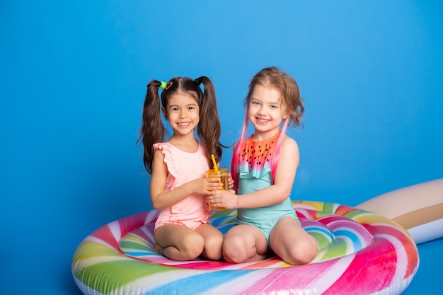 Una ragazza felice due del bambino in costume da bagno che beve il succo d'arancia che si siede sul materasso gonfiabile variopinto sulla superficie blu.