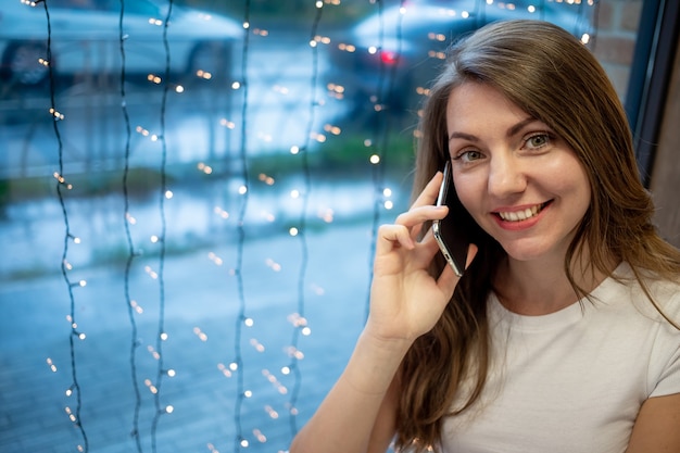 Una ragazza freelance sorridente che lavora a un laptop in un bar e parla al telefono, la giornata lavorativa di una giovane donna o studente libera