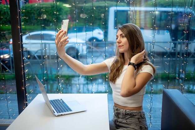 Una ragazza freelance sorridente si fa un selfie al telefono o fa una videochiamata, una giornata lavorativa di una giovane donna libera o di uno studente con un laptop