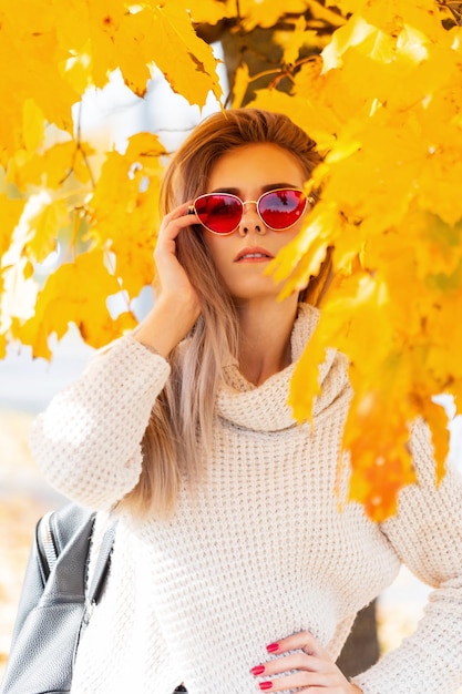 Una ragazza fresca ed elegante con occhiali eleganti in un maglione bianco vintage con zaino si trova nel luminoso fogliame autunnale dorato nel parco