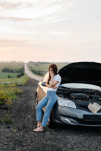 Una ragazza frustrata si trova vicino a un'auto in panne nel mezzo dell'autostrada durante il tramonto Guasto e riparazione dell'auto In attesa di aiuto Servizio auto Guasto auto su strada