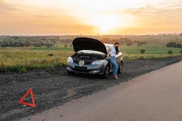 Una ragazza frustrata si trova vicino a un'auto in panne nel mezzo dell'autostrada durante il tramonto Guasto e riparazione dell'auto In attesa di aiuto Servizio auto Guasto auto su strada