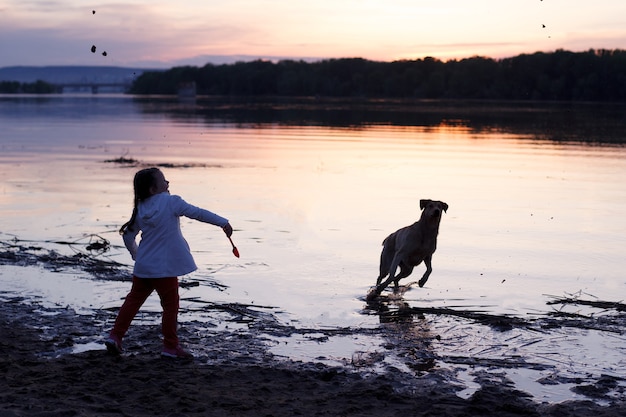 Una ragazza gioca con un cane su una spiaggia di sabbia in riva al fiume al tramonto.