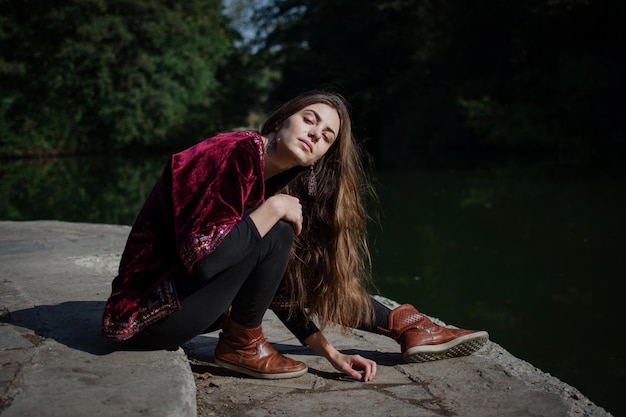 Una ragazza giovane e libera in abiti esotici si siede sulla riva del fiume e ti guarda