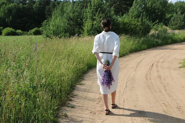 Una ragazza in abiti bianchi cammina lungo una strada di campagna con un mazzo di fiori di campo alle spalle