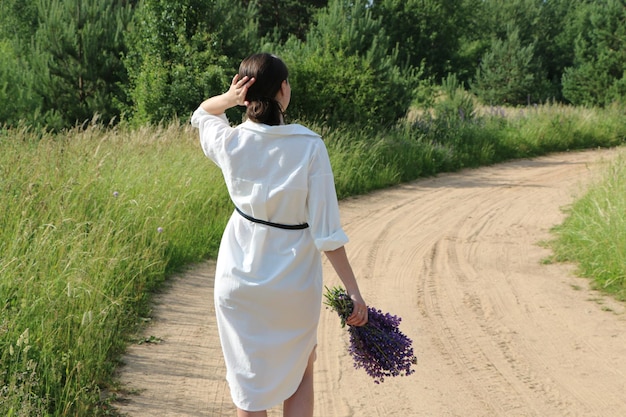 Una ragazza in abiti bianchi cammina lungo una strada di campagna con un mazzo di fiori di campo