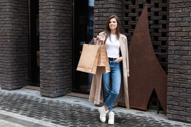 Una ragazza in abiti casual con borse della spesa e molti acquisti vicino al centro commerciale ti consegna sorridente il suo pacchetto consumismo vendita e concetto di persone