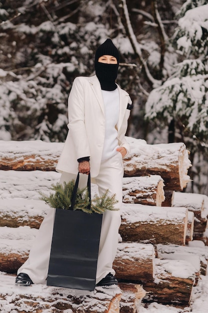 Una ragazza in abito bianco e passamontagna con un pacchetto di alberi di Natale nella foresta invernale a CapodannoConcetto di Capodanno