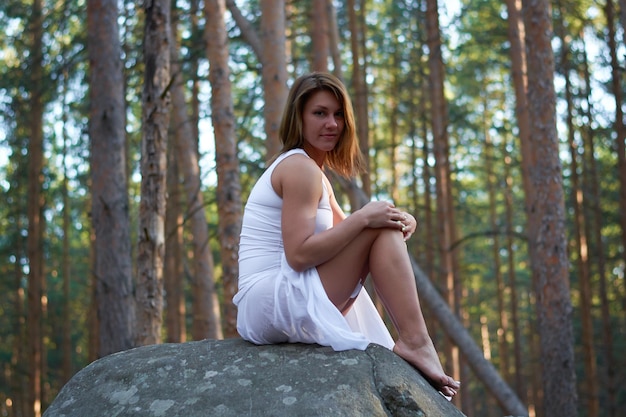 Una ragazza in abito bianco si siede su una roccia nella foresta al tramonto