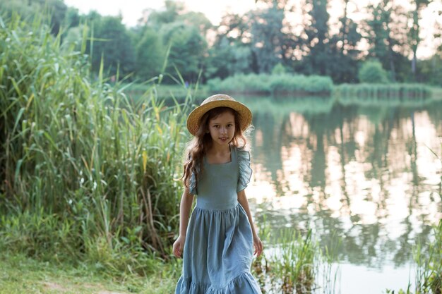 Una ragazza in abito blu e cappello di paglia si trova sulla riva del lago