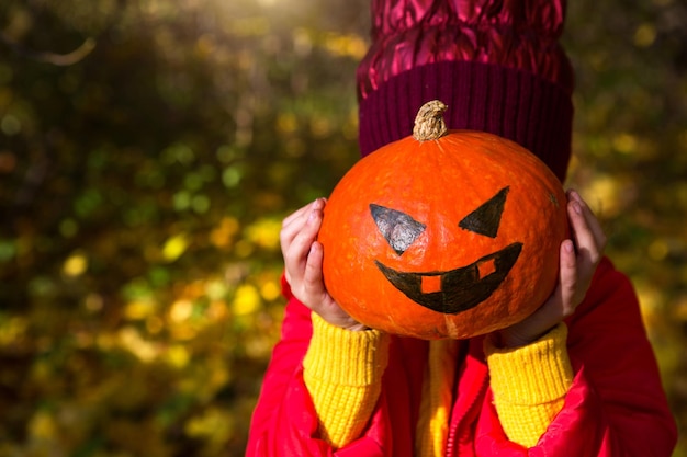 Una ragazza in caldi abiti rossi e gialli tiene una zucca con occhi dipinti e bocca nelle sue mani Halloween Jack Lantern senza una faccia Umore autunnale giallo foglie secche