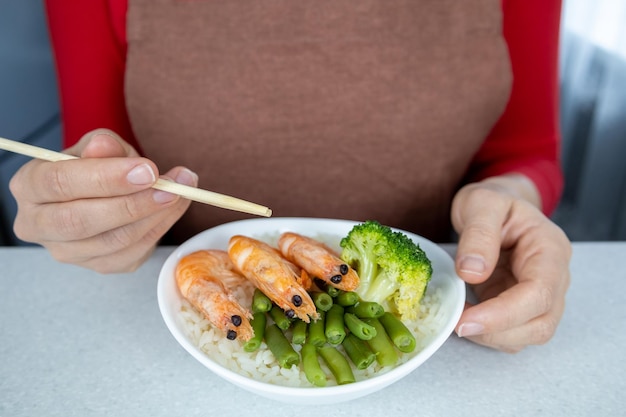 Una ragazza in camicetta rossa e grembiule marrone tiene in mano un piatto di riso dorato e tre grandi gamberoni Il piatto è decorato con fagiolini e broccoli Concetto di cibo asiatico