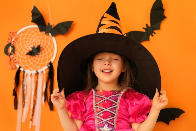 Una ragazza in costume da strega su uno sfondo arancione con pipistrelli tiene un cappello che chiude sognante gli occhi