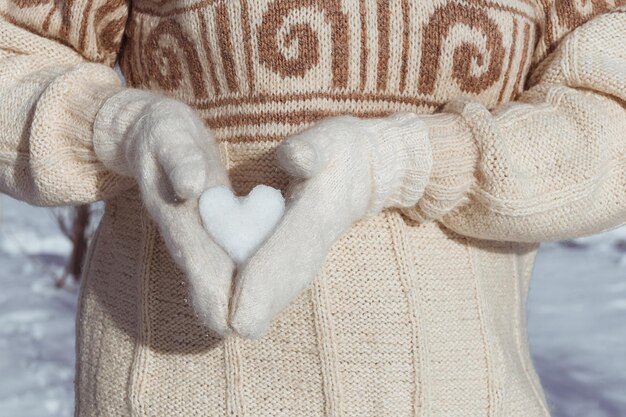 Una ragazza in maglione beige tiene il cuore fatto di neve nelle mani vestite di guanti per San Valentino