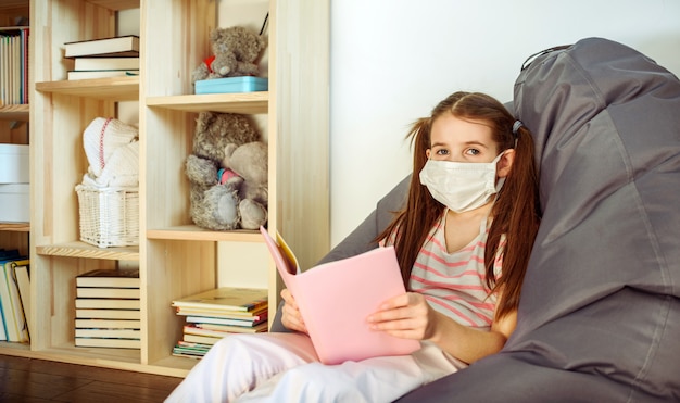 Una ragazza in maschera medica ubicazione sul pavimento di legno e la lettura di libri durante la quarantena