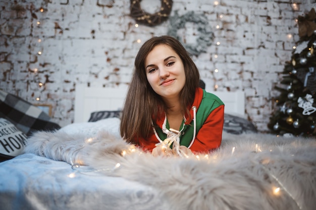 Una ragazza in tuta di Capodanno con un umore festoso giace su un letto attorno a ghirlande luminose