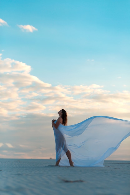 Una ragazza in un abito bianco vola balla e pone nel deserto di sabbia al tramonto