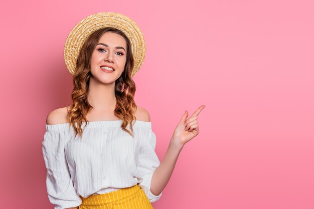 Una ragazza in un cappello di paglia e un abito vintage bianco sorride e punta un dito verso. La ragazza in estate copre su un dito rosa di manifestazione della parete