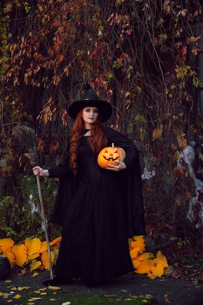 una ragazza in un costume da strega nera e un cappello a punta con una scopa e una zucca in un parco ad Halloween