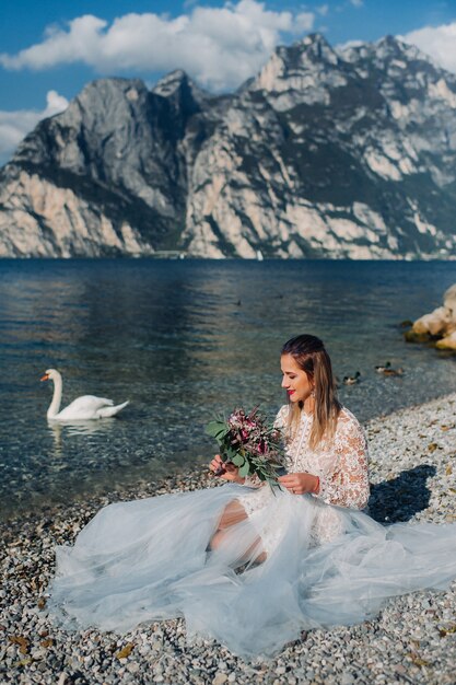 Una ragazza in un elegante abito bianco è seduta sull'argine del lago di Garda.Una donna è fotografata sullo sfondo di una montagna e di un lago in Italia.Torbole