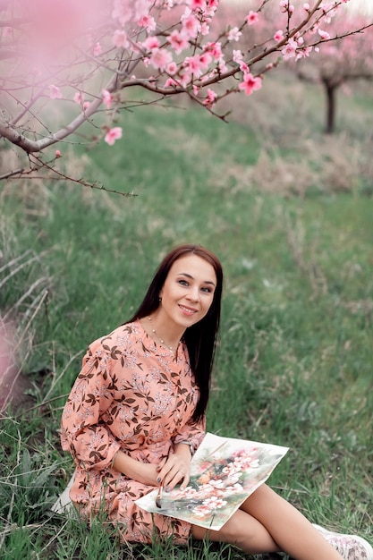 Una ragazza in un frutteto di pesche in fiore sta riposando sotto un albero seduto e disegnando