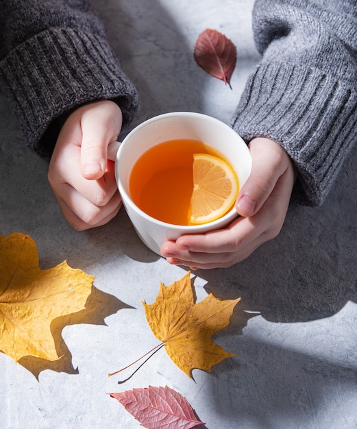 Una ragazza in un maglione grigio tiene una tazza di tè con il limone in mano su sfondo grigio. luce del mattino. Vista frontale e da vicino