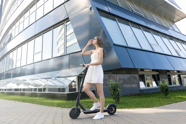 Una ragazza in un vestito cavalca uno scooter elettrico vicino a edifici moderni