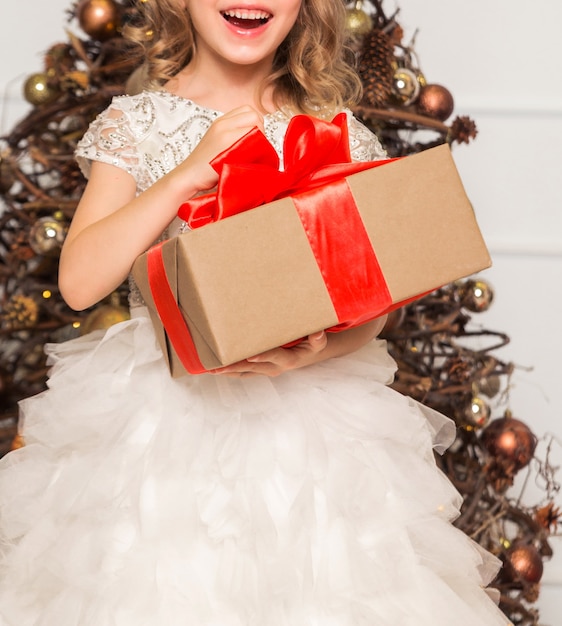 Una ragazza in un vestito chic sta con un regalo di Capodanno tra le mani sullo sfondo di un albero di Capodanno