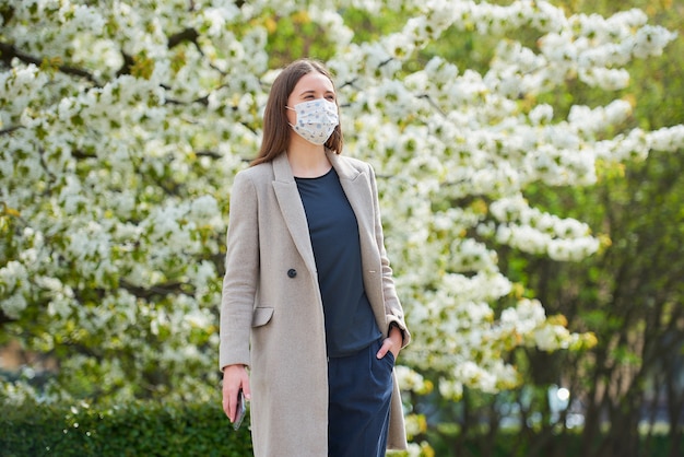 Una ragazza in una maschera medica per evitare la diffusione del coronavirus tiene uno smartphone nel parco. Una donna con una maschera contro COVID-19 mantiene una distanza sociale nel giardino tra alberi in fiore.