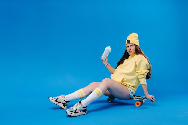 Una ragazza incinta in abiti gialli con un bicchiere di succo si siede su uno skateboard su uno sfondo blu