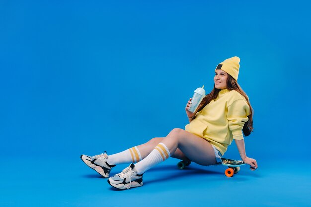 Una ragazza incinta in abiti gialli con un bicchiere di succo si siede su uno skateboard su uno sfondo blu.