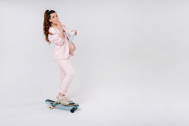 Una ragazza incinta in un abito rosa con uno skateboard in mano si trova su uno sfondo grigio