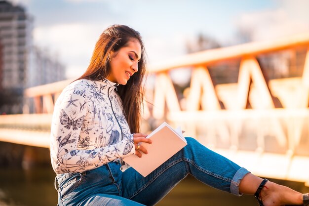 Una ragazza latina bruna in città al tramonto in riva al fiume che si diverte a leggere un libro