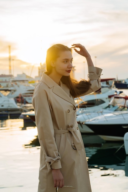 Una ragazza lussuosa ed elegante con un cappotto beige sta aspettando il suo yacht nel porto di un'auto al tramonto