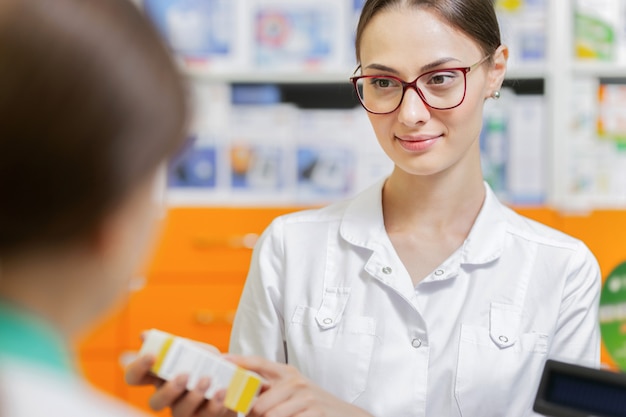 Una ragazza magra e amichevole con i capelli scuri e gli occhiali, che indossa un camice da laboratorio, suggerisce le pillole a un visitatore alla cassa di una farmacia. .