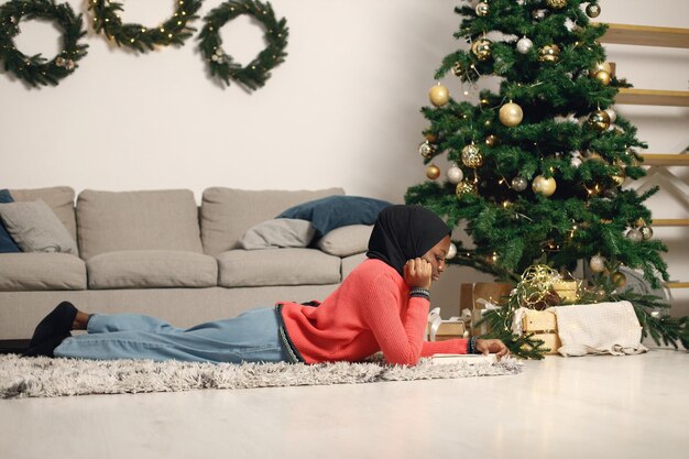 Una ragazza musulmana in hijab nero giaceva vicino all'albero di Natale con un libro