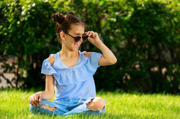 Una ragazza nel parco in abiti blu seduta sull'erba. un bambino con gli occhiali da sole. Foto di alta qualità