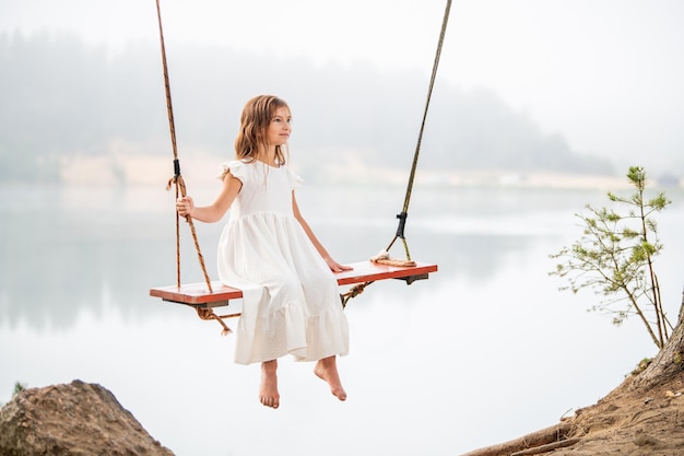 Una ragazza nella foresta che oscilla su un'altalena Altalena di corda su un lago nella foresta Ragazza a piedi nudi con un abito bianco e capelli lunghi