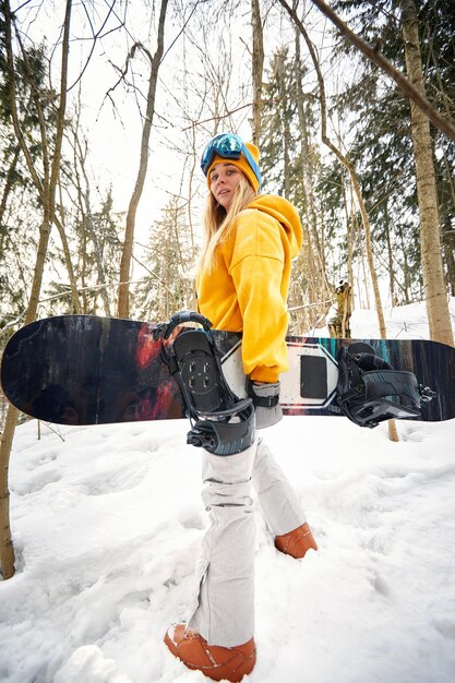 Una ragazza o una donna snowboarder si dedica agli sport invernali nella foresta innevata, sta nella neve e tiene in mano uno snowboard