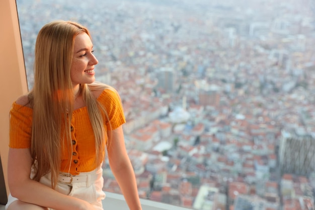 Una ragazza pensierosa siede vicino a una finestra panoramica su un piano alto di un edificio e guarda la città dal cielo