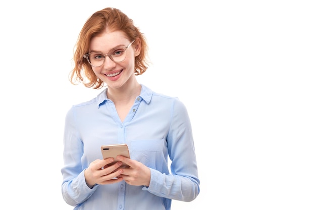 Una ragazza rossa sorridente felice che tiene in mano lo smartphone con gli occhiali e la camicia da lavoro isolata su sfondo bianco per studio, ha ricevuto un messaggio gioioso