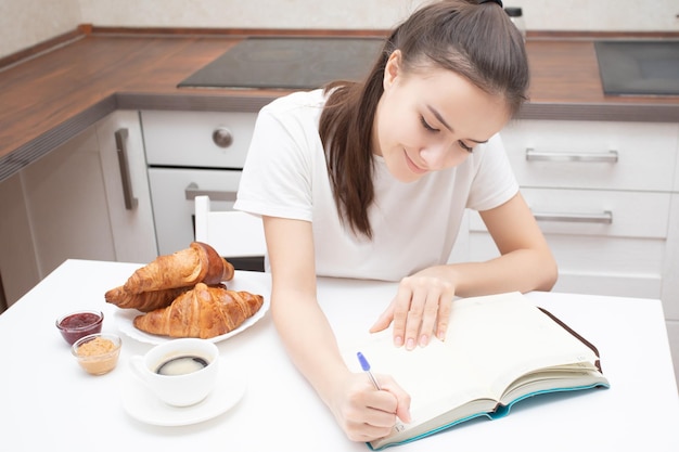 Una ragazza scrive un programma in un quaderno e pianifica il suo calendario al mattino in cucina per la colazione