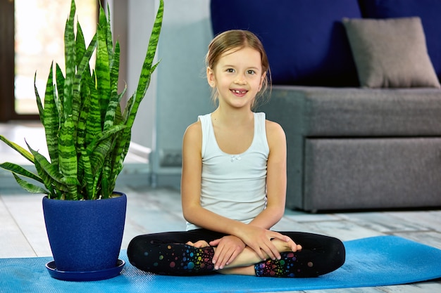 Una ragazza seduta in asana yoga su una superficie blu. Il concetto di formazione domestica, yoga per bambini.