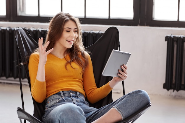 Una ragazza si siede a casa su un'elegante sedia in pelle nera, comunica tramite videochiamata su un tablet. Lavoro e relax a casa