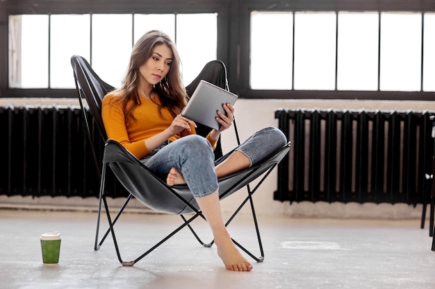 Una ragazza si siede a casa su un'elegante sedia in pelle nera, tiene in mano una tazza di caffè e un tablet. Lavoro e relax a casa