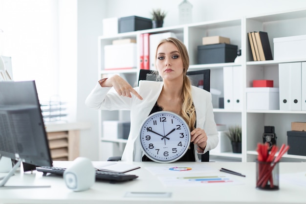 Una ragazza si siede a un tavolo in ufficio e punta un dito sul quadrante dell'orologio.