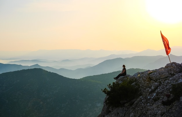 Una ragazza si siede in montagna sullo sfondo del tramonto vicino alla bandiera rossa