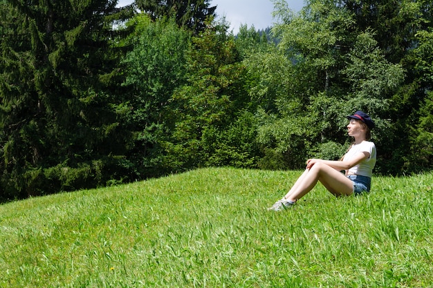 Una ragazza si siede su uno sfondo di montagne sul prato verde e gode del sole.